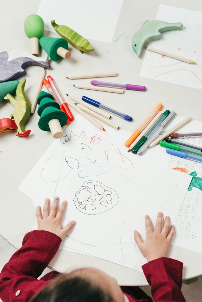 A child engages in creative drawing with colored pencils surrounded by wooden toys on a table.